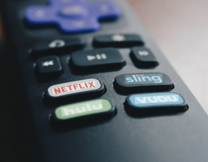 A Roku remote placed on a coffee table next to a smart TV displaying a streaming home screen.